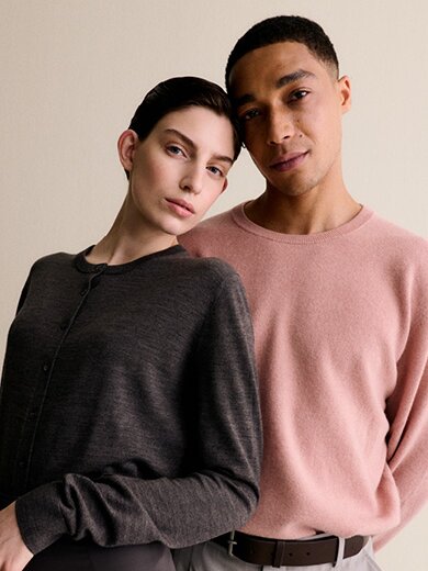 Eine Frau in einem grauen Pullover und ein Mann in einem rosafarbenen Pullover lehnen vor einem hellen Hintergrund aneinander und wirken entspannt. | © Gabor Shoes GmbH, Rosenheim