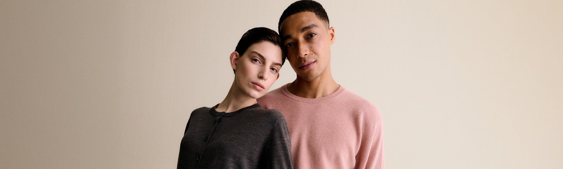 Eine Frau in einem grauen Pullover und ein Mann in einem rosafarbenen Pullover lehnen vor einem hellen Hintergrund aneinander und wirken entspannt. | © Gabor Shoes GmbH, Rosenheim