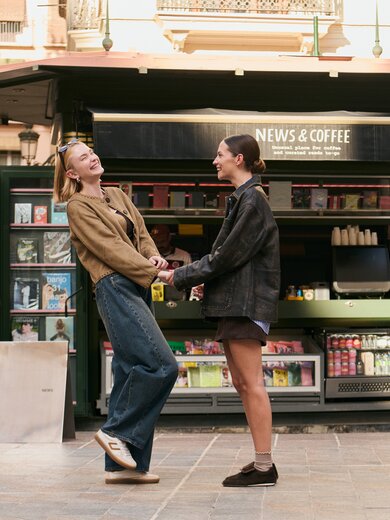 Due donne stanno davanti a un chiosco aperto con la scritta "News & Coffee", si tengono per mano e si sorridono. Sullo sfondo si vedono riviste e bevande. | © Gabor Shoes GmbH, Germania