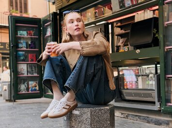 Una donna è seduta rilassata su un blocco di granito davanti a un chiosco aperto, tiene in mano un bicchiere e indossa sneaker Gabor chiare, jeans blu e una giacca beige. Sullo sfondo si vedono bevande e riviste. | © Gabor Shoes GmbH, Germania