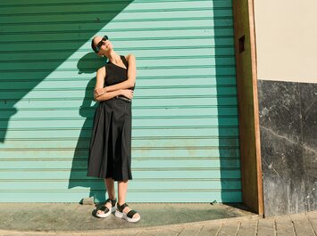 Una donna sta rilassata con le braccia incrociate davanti a una porta del garage turchese. Indossa occhiali da sole neri, un top nero senza maniche, pantaloni neri ampi e sandali neri con suola bianca. | © Gabor Shoes GmbH, Germania