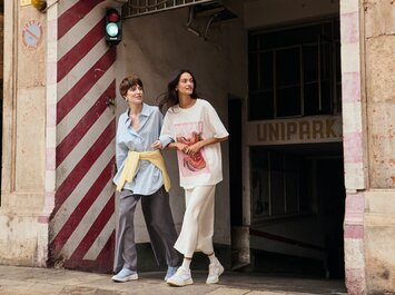 Due donne escono a braccetto da un parcheggio sotterraneo sul marciapiede, accanto a un muro con strisce rosse e bianche e un semaforo verde. Entrambe indossano abiti casual e sneaker chiare. | © Gabor Shoes GmbH, Germania
