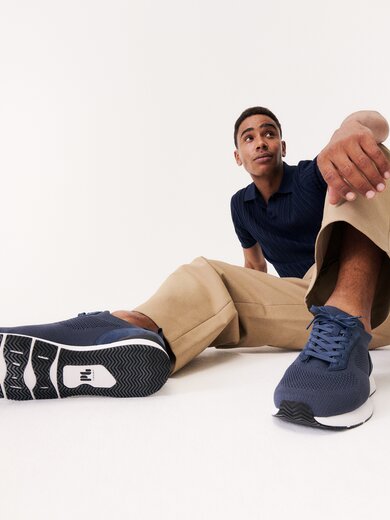 Un uomo è seduto in modo rilassato sul pavimento, indossa pantaloni beige, una maglia blu e sneakers blu scuro. Le scarpe sono ben visibili in primo piano nell’immagine. | © Gabor Shoes GmbH, Germania