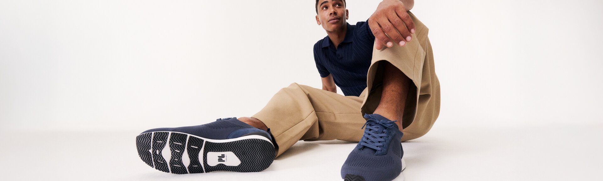 Un uomo è seduto in modo rilassato sul pavimento, indossa pantaloni beige, una maglia blu e sneakers blu scuro. Le scarpe sono ben visibili in primo piano nell’immagine. | © Gabor Shoes GmbH, Germania