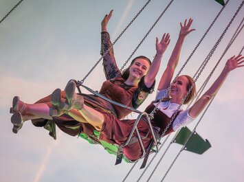 Zwei Frauen sitzen nebeneinander auf einem Kettenkarussell und strecken fröhlich die Arme in die Luft. Sie tragen traditionelle Dirndl und das Bild zeigt den Himmel im Hintergrund. | © Gabor Shoes GmbH, Rosenheim