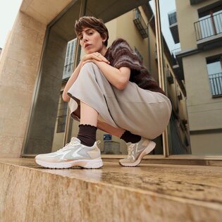 Eine Frau hockt auf einer steinernen Stufe vor einem modernen Gebäude und trägt helle Sneakers, dunkle Socken, einen weiten Rock und einen kurzärmeligen Pullover. | © Gabor Shoes GmbH, Rosenheim