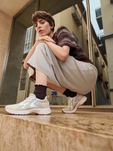 Una donna è accovacciata su un gradino di pietra davanti a un edificio moderno e indossa sneakers chiare, calze scure, una gonna ampia e un maglione a maniche corte. | © Gabor Shoes GmbH, Germania