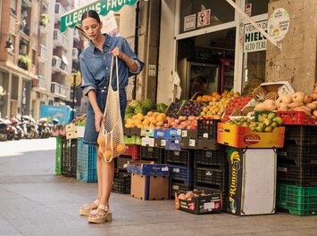 Una donna si trova davanti a una bancarella di frutta e verdura all’aperto e tiene una rete di arance. Indossa un vestito di jeans blu e sandali chiari. Sullo sfondo si vedono molte cassette colorate di frutta e verdura e una scena di strada. | © Gabor Shoes GmbH, Germania