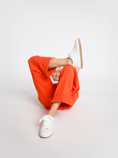 Eine Frau mit oranger Hose und weißen Sneakern sitzt auf dem Boden, ein Bein angewinkelt, im hellen, minimalistischen Raum. | © Gabor Shoes GmbH, Rosenheim