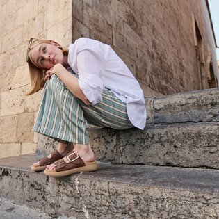 Eine Frau sitzt auf einer steinernen Treppe vor einer alten Mauer. Sie trägt eine weiße Bluse, weit geschnittene, gestreifte Hose in Grün und Beige sowie braune Sandalen mit goldener Schnalle. Sie hat sich eine Sonnenbrille in die Haare geschoben.  | © Gabor Shoes GmbH, Rosenheim