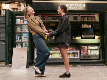 Zwei Frauen stehen vor einem offenen Kiosk mit der Aufschrift "News & Coffee", halten sich an den Händen und lächeln sich an. Im Hintergrund sind Zeitschriften und Getränke zu sehen. | © Gabor Shoes GmbH, Rosenheim