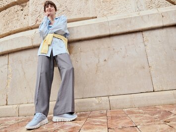 Una donna è in piedi davanti a un muro di pietra su piastrelle con motivi, indossa una camicetta azzurra, un cardigan giallo legato in vita, pantaloni grigi ampi e sneaker azzurri. La foto è scattata dal basso (prospettiva dal basso). | © Gabor Shoes GmbH, Germania