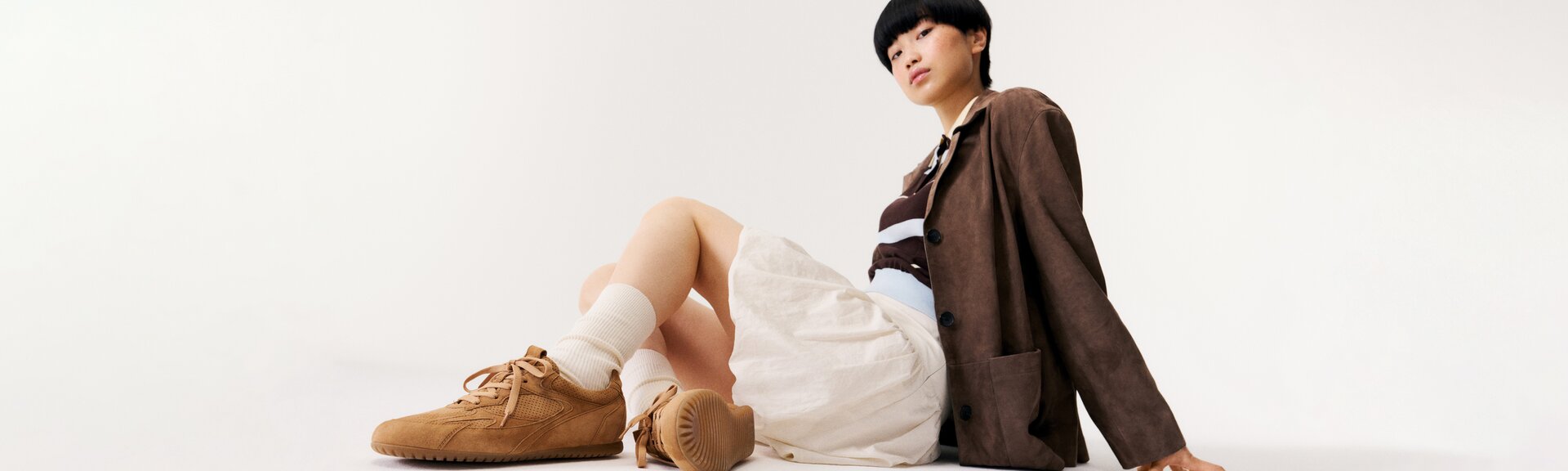 Una donna è seduta per terra su uno sfondo bianco e indossa sneaker marroni, calzini bianchi, una gonna beige, una maglia a righe e un blazer marrone. | © Gabor Shoes GmbH, Germania