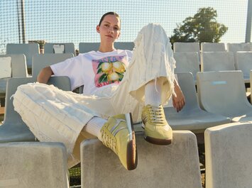 Una donna è seduta per terra in una stanza bianca e indossa sneaker marroni, calzini bianchi, una gonna beige, una maglia a righe e un blazer marrone. | © Gabor Shoes GmbH, Germania