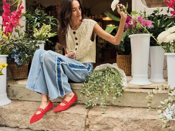 Una donna è seduta su una scalinata di pietra davanti a un negozio di fiori, circondata da fiori colorati e vasi bianchi. Indossa un gilet chiaro in maglia, jeans larghi e scarpe rosse vistose. In mano tiene uno stelo di fiore. | © Gabor Shoes GmbH, Germania