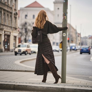 Eine Frau steht in herbstlicher Natur auf einem Kiesweg, trägt braune Stiefel, eine braune Jacke und einen kurzen Rock, und lehnt sich an trockenes, hohes Schilfgras. | © Gabor Shoes GmbH, Rosenheim