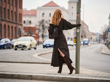 Eine Frau steht in herbstlicher Natur auf einem Kiesweg, trägt braune Stiefel, eine braune Jacke und einen kurzen Rock, und lehnt sich an trockenes, hohes Schilfgras. | © Gabor Shoes GmbH, Rosenheim
