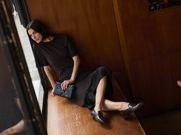 Eine Frau mit lockigen Haaren lehnt an einer hellen Steinwand, trägt ein langes schwarzes Kleid und braune Stiefel. Die Szene ist sonnig und ruhig. | © Gabor Shoes GmbH, Rosenheim