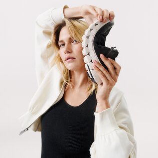 Eine Frau hält einen flexiblen, schwarzen Sneaker mit weißer Sohle neben ihren Kopf. Sie trägt eine weiße Jacke und ein schwarzes Oberteil vor hellem Hintergrund. | © Gabor Shoes GmbH, Rosenheim