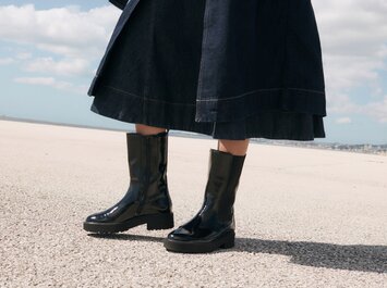 Eine Frau steht draußen auf hellem Boden und trägt einen dunklen Rock sowie glänzende schwarze Stiefel. Der Himmel ist blau mit einigen Wolken. | © Gabor Shoes GmbH, Rosenheim