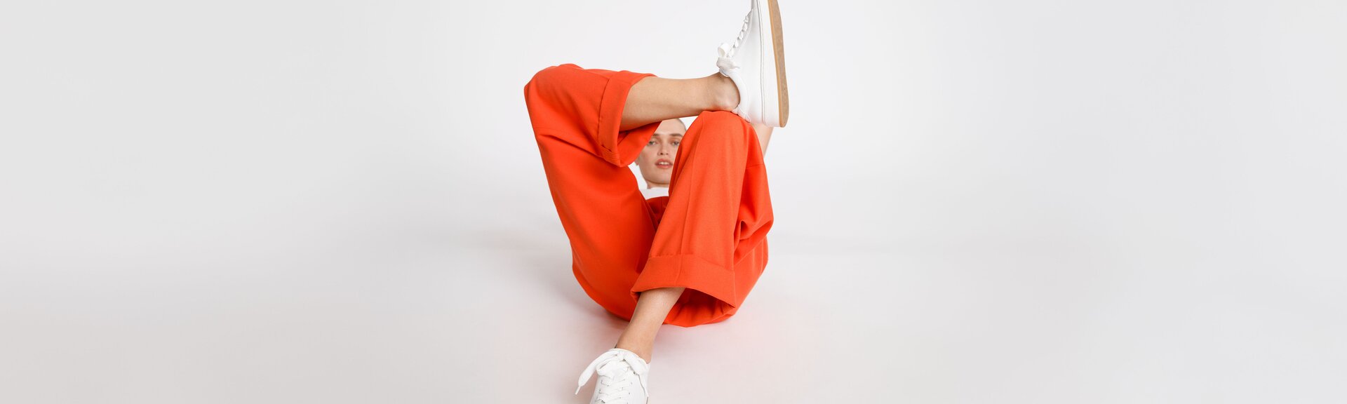 Eine Frau mit oranger Hose und weißen Sneakern sitzt auf dem Boden, ein Bein angewinkelt, im hellen, minimalistischen Raum. | © Gabor Shoes GmbH, Rosenheim
