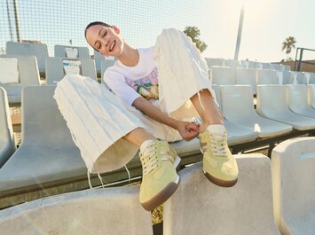 Eine Frau mit ausgestellten, weißen Hosen und gelben Sneakern sitzt auf leeren Stadion-Sitzen in der Sonne und schlüpft in ihren linken Schuh. | © Gabor Shoes GmbH, Rosenheim