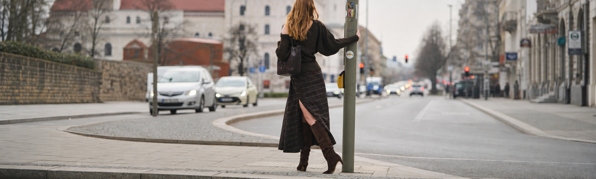 Eine Frau steht in herbstlicher Natur auf einem Kiesweg, trägt braune Stiefel, eine braune Jacke und einen kurzen Rock, und lehnt sich an trockenes, hohes Schilfgras. | © Gabor Shoes GmbH, Rosenheim