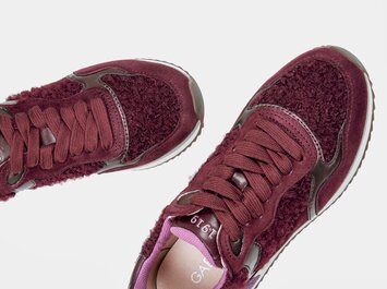 Eine Frau mit beigefarbener Hose trägt bordeauxrote Sneaker und steht vor zwei Spiegeln, sodass die Schuhe mehrfach reflektiert werden. | © Gabor Shoes GmbH, Rosenheim
