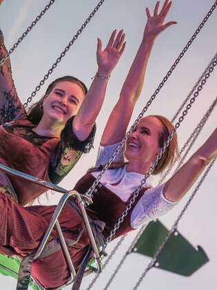Zwei Frauen sitzen nebeneinander auf einem Kettenkarussell und strecken fröhlich die Arme in die Luft. Sie tragen traditionelle Dirndl und das Bild zeigt den Himmel im Hintergrund. | © Gabor Shoes GmbH, Rosenheim