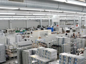 Große Produktionshalle mit Regalen, Kartons, Maschinen und mehreren arbeitenden Personen.