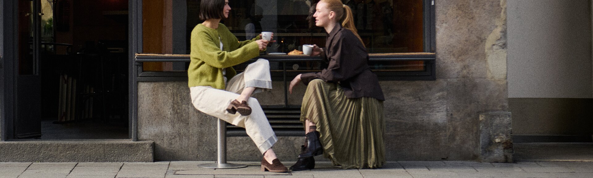 Eine Frau sitzt in einem hellen Raum, trägt ein lilafarbenes Kleid und einen schwarzen Blazer sowie schwarze Sandalen mit Riemen und dekorativen Details. | © Gabor Shoes GmbH, Rosenheim