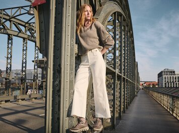 Eine Frau steht lässig vor einer hellen Steinwand, trägt weite graue Hose, hellblaue Sneaker, ein langes, blau gestreiftes Hemd und hat einen gelben Pullover locker um die Hüfte gebunden. Fotografiert aus Frosch-Perspektive. | © Gabor Shoes GmbH, Rosenheim