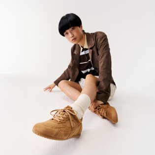 Eine Frau sitzt auf dem Boden in einem weißen Raum und trägt braune Sneaker, weiße Socken, einen beigen Rock, ein gestreiftes Oberteil und einen braunen Blazer. | © Gabor Shoes GmbH, Rosenheim