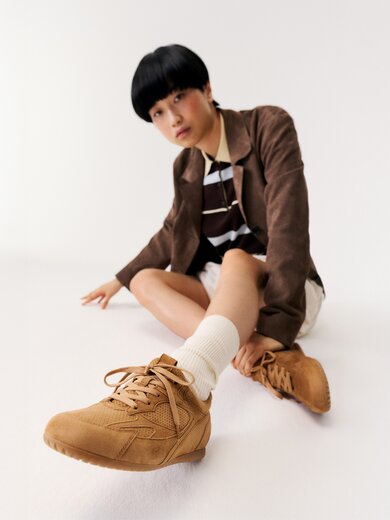 Una donna è seduta per terra in una stanza bianca e indossa sneaker marroni, calzini bianchi, una gonna beige, una maglia a righe e un blazer marrone. | © Gabor Shoes GmbH, Germania