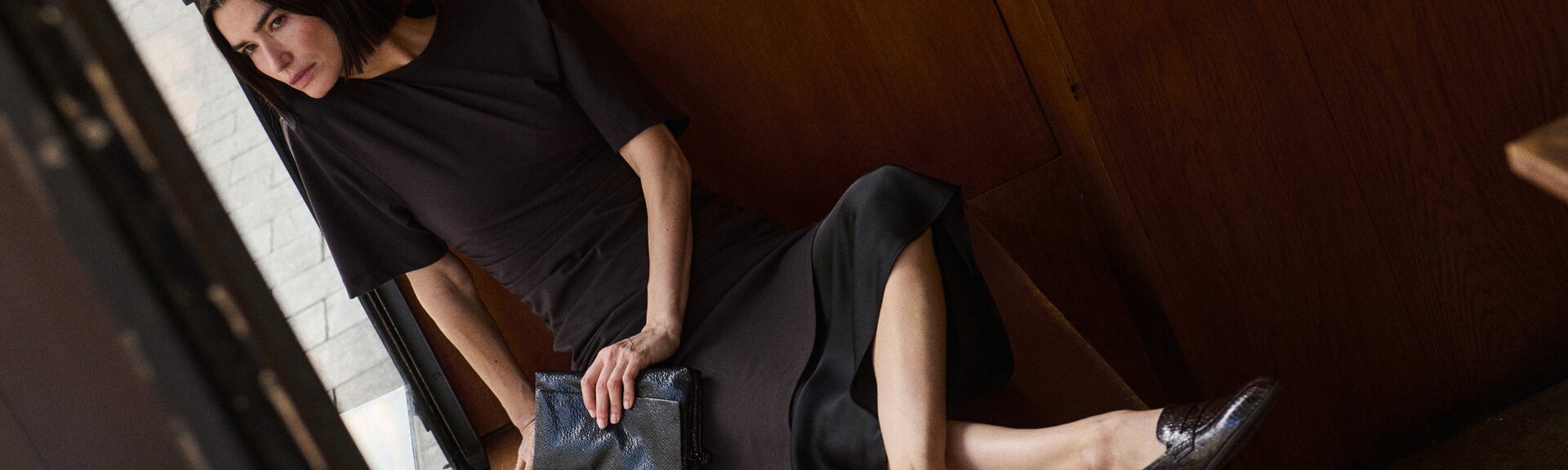 Eine Frau steht entspannt an einer hellen Steinwand, trägt ein dunkles langes Kleid und braune Stiefel. Das Bild ist bei Tageslicht im Freien aufgenommen. | © Gabor Shoes GmbH, Rosenheim