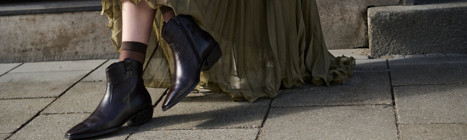 Eine Frau trägt dunkelbraune Damen-Stiefeletten mit spitzer Kappe und kleinem Absatz zu einem langen, fließenden olivgrünen Rock; sie steht auf einem gepflasterten Gehweg vor Steinstufen. | © Gabor Shoes GmbH, Rosenheim