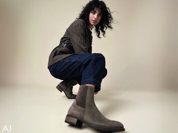 Ein mit KI generiertes Bild einer Frau, die vor hellem Hintergrund hockt und eine braune Jacke, dunkelblaue Jeans und Gabor-Chelsea-Boots trägt.  | © Gabor Shoes GmbH, Rosenheim