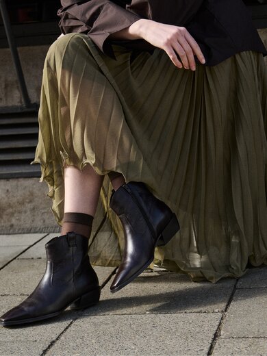Eine Frau trägt dunkelbraune Damen-Stiefeletten mit spitzer Kappe und kleinem Absatz zu einem langen, fließenden olivgrünen Rock; sie steht auf einem gepflasterten Gehweg vor Steinstufen. | © Gabor Shoes GmbH, Rosenheim