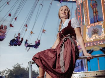Eine Frau in einem traditionellen bayerischen Dirndl sitzt auf einem Geländer vor einem farbenfrohen Kettenkarussell auf einem Volksfest. Im Hintergrund sind Fahrgäste in Bewegung und das Karussell ist mit bunten Lichtern dekoriert. | © Gabor Shoes GmbH, Rosenheim