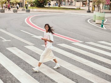Una donna attraversa una strada larga sulle strisce pedonali. Indossa una t-shirt bianca, una gonna lunga chiara e sneakers bianche, e tiene uno smartphone in mano. Sullo sfondo si vedono alberi e alcuni passanti. | © Gabor Shoes GmbH, Germania