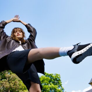 Eine Frau steht im Freien, trägt eine dunkle Jacke, schwarzen Rock und schwarze Sneaker, ein Bein ist in die Luft gestreckt. | © Gabor Shoes GmbH, Rosenheim