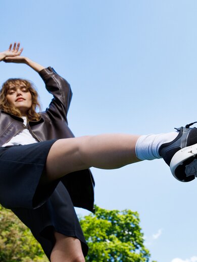 Eine Frau steht im Freien, trägt eine dunkle Jacke, schwarzen Rock und schwarze Sneaker, ein Bein ist in die Luft gestreckt. | © Gabor Shoes GmbH, Rosenheim