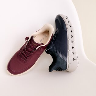 Eine Frau sitzt auf dem Boden in einem hellen Raum und trägt rosa Sneaker, gestreifte Socken, einen cremefarbenen Rock und eine rosa Jacke. | © Gabor Shoes GmbH, Rosenheim