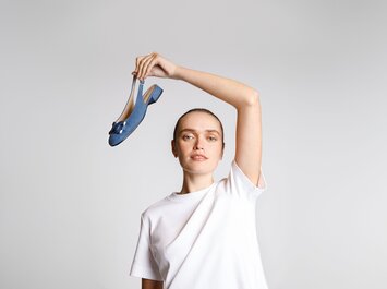 Eine Frau in weißem T-Shirt hält einen blauen Schuh mit kleinem Absatz und Schleife hoch in die Luft, vor hellem Hintergrund. | © Gabor Shoes GmbH, Rosenheim