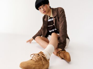 Eine Frau sitzt auf dem Boden in einem weißen Raum und trägt braune Sneaker, weiße Socken, einen beigen Rock, ein gestreiftes Oberteil und einen braunen Blazer. | © Gabor Shoes GmbH, Rosenheim