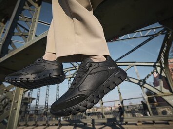 Ein Mann sitzt auf einer Stahlbrücke, trägt hellbeige Hose und schwarze Gabor-Sneaker, seine Füße hängen über der Straße. | © Gabor Shoes GmbH, Rosenheim