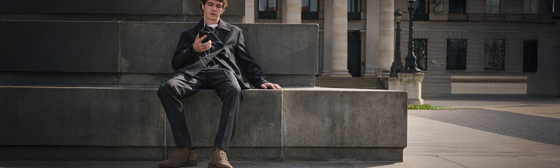Junger Mann sitzt entspannt auf einer Steinbank, hört mit Kopfhörern Musik auf seinem Smartphone und trägt einen dunkelgrauen Anzug mit braunen Gabor-Schnürboots. | © Gabor Shoes GmbH, Rosenheim