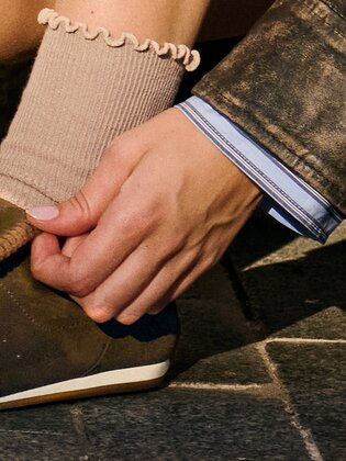 Eine Person kniet auf einem gepflasterten Gehweg und zieht ihren braunen Sneaker mit beigem Socken an. | © Gabor Shoes GmbH, Rosenheim