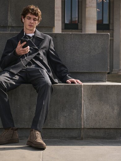Junger Mann sitzt entspannt auf einer Steinbank, hört mit Kopfhörern Musik auf seinem Smartphone und trägt einen dunkelgrauen Anzug mit braunen Gabor-Schnürboots. | © Gabor Shoes GmbH, Rosenheim