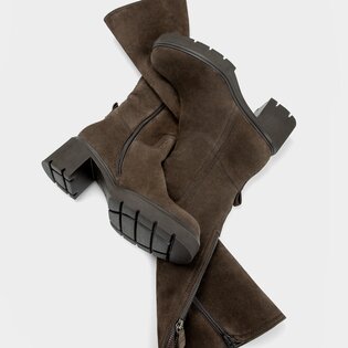 "Eine Frau sitzt entspannt in eleganter brauner Kleidung und braunen Stiefeln auf sandigem Boden zwischen Dünen und Gräsern. Im Hintergrund sind grüne Büsche und Bäume zu sehen. " | © Gabor Shoes GmbH, Rosenheim